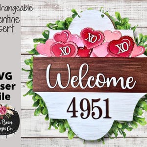 May include: Interchangeable Valentine's Day insert for a welcome sign. The insert features a white background with a brown wooden frame and the text "Welcome" in white. The insert also features a white background with a brown wooden frame and the number "4951" in black. The insert is surrounded by a green leafy garland with red and pink hearts with "XO" written on them.