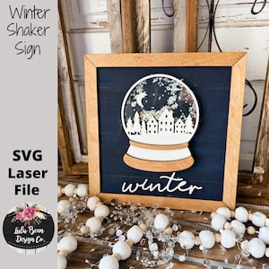 May include: Wooden winter shaker sign with a snow globe design. The sign features a wooden frame, a navy blue background, and a white snow globe with a winter scene. The word "winter" is written in white script.