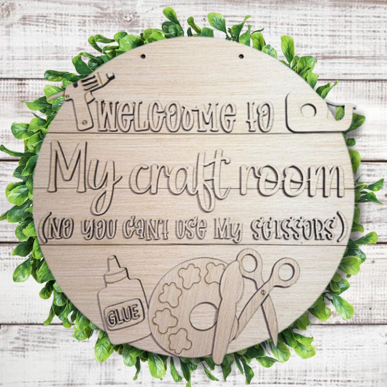 May include: A round wooden sign with the text "Welcome to My craft room (No you can't use My scissors)". The sign features a glue bottle, paint palette, and scissors. The sign is surrounded by green leaves.