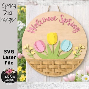 May include: A round wooden door hanger with the words "Welcome Spring" in pink. The design features three tulips in pink, yellow, and blue, set in a green basket. The words "Spring Door Hanger" and "SVG Laser File" are also visible.