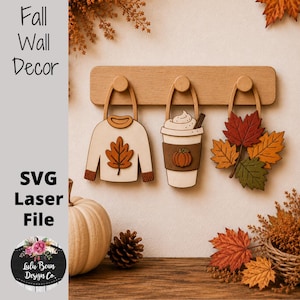 Peut inclure: Un porte-manteau mural en bois clair avec trois crochets supportant des décorations suspendues : un pull crème avec une feuille orange, un gobelet à café avec un motif de citrouille et de la crème fouettée, et des feuilles d'automne.