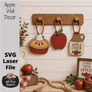 Puede incluir: Un soporte de pared de madera con tres adornos colgantes: un pastel con nata montada, una manzana roja y una jarra color canela etiquetada como Apple Cider. Los adornos cuelgan de una tabla horizontal. Incluye un letrero que dice Farm Fresh Apples.