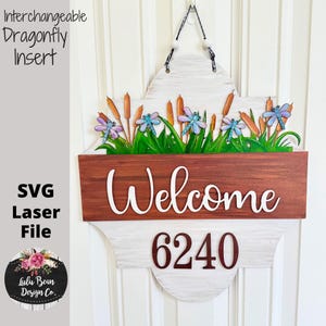 May include: Wooden welcome sign with interchangeable dragonfly insert. The sign features a brown wooden section with the word "Welcome" in white script, and the number "6240". The top section has a white distressed finish, and the sign is decorated with dragonflies, cattails, and green grass.