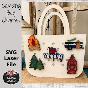 May include: A white tote bag cutout with circular holes, featuring five decorative camping-themed charms: a campfire, a blue and white camper, two green pine trees, a red lantern, and a blue sign with the text I love camping.