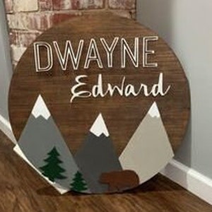 K&ouml;nnte beinhalten: Ein rundes Holzschild mit den Namen "DWAYNE" und "Edward", die in Wei&szlig; bemalt sind. Das Schild zeigt eine Berglandschaft mit grauen und braunen Bergen, gr&uuml;nen B&auml;umen und einem braunen B&auml;ren.