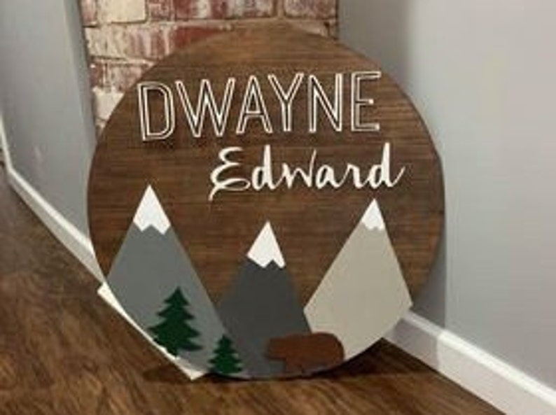 K&ouml;nnte beinhalten: Ein rundes Holzschild mit den Namen "DWAYNE" und "Edward", die in Wei&szlig; bemalt sind. Das Schild zeigt eine Berglandschaft mit grauen und braunen Bergen, gr&uuml;nen B&auml;umen und einem braunen B&auml;ren.