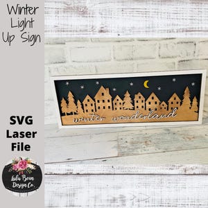 May include: A wooden sign with a white frame features a winter wonderland scene. The design includes silhouettes of houses, trees, stars, and a crescent moon against a dark background. The words "winter wonderland" are written in cursive.