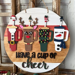 Pode incluir: Placa redonda de madeira com quatro desenhos festivos de x&iacute;caras de caf&eacute;: Papai Noel, rena, luzes de Natal e um boneco de neve. A placa diz "Have a cup of cheer" em letras pretas e verdes. Uma corda de juta &eacute; anexada para pendurar.