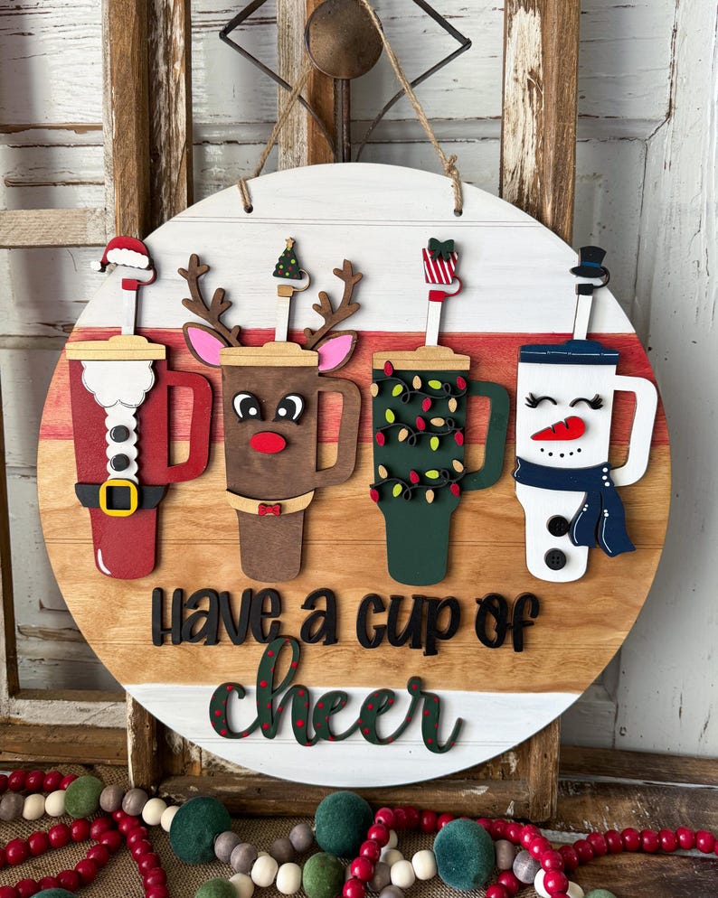Pode incluir: Placa redonda de madeira com quatro desenhos festivos de x&iacute;caras de caf&eacute;: Papai Noel, rena, luzes de Natal e um boneco de neve. A placa diz "Have a cup of cheer" em letras pretas e verdes. Uma corda de juta &eacute; anexada para pendurar.
