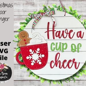 May include: A white round wooden door hanger with a green leafy wreath around the edge. The center of the hanger has a red mug with a white snowflake and a gingerbread man. The text "Have a cup of cheer" is written in red and green letters.