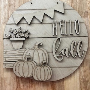 Puede incluir: Un letrero redondo de madera con las palabras "Hello Fall" en una fuente decorativa. El letrero tambi&eacute;n presenta una pancarta, una planta en maceta y tres calabazas.