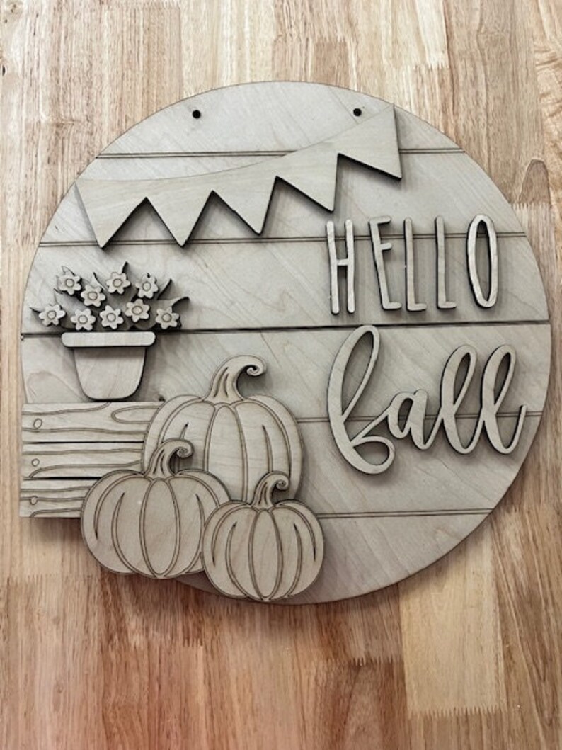 Puede incluir: Un letrero redondo de madera con las palabras "Hello Fall" en una fuente decorativa. El letrero tambi&eacute;n presenta una pancarta, una planta en maceta y tres calabazas.