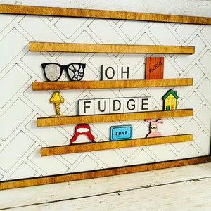 May include: A wooden framed wall decor with a white background and a black and white diamond pattern. The frame has four shelves with various items on them, including a pair of glasses, a lamp, the word "OH FUDGE" spelled out in Scrabble tiles, a house, a red hat, a blue bar of soap, and a pink pig in a pink sweater.