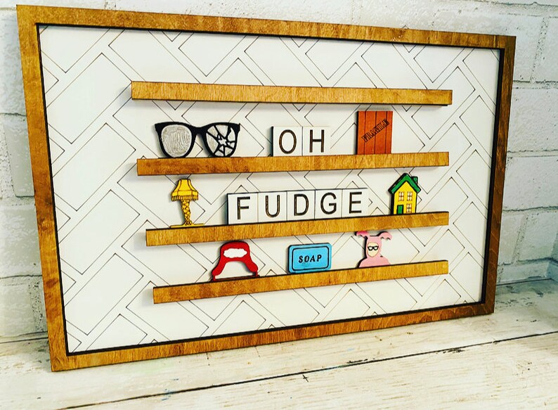 May include: A wooden framed wall decor with a white background and a black and white diamond pattern. The frame has four shelves with various items on them, including a pair of glasses, a lamp, the word "OH FUDGE" spelled out in Scrabble tiles, a house, a red hat, a blue bar of soap, and a pink pig in a pink sweater.