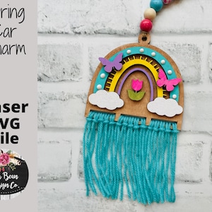 May include: A wooden rainbow charm with a blue, yellow, and white color scheme. The rainbow has a pink flower in the center and two purple butterflies on either side. The charm is attached to a blue tassel.