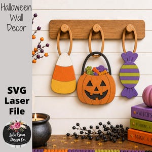 May include: A wooden wall rack with three hanging decorations: a candy corn, a jack-o'-lantern bucket filled with colorful candies, and a purple and green striped wrapped candy. The rack is mounted on a white horizontal paneled wall.