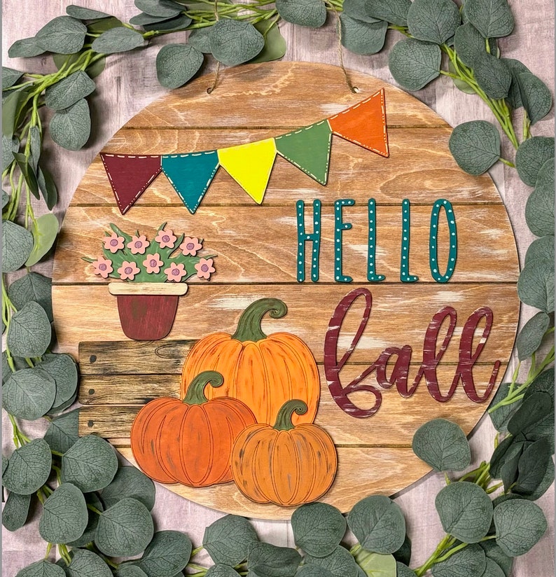 Puede incluir: Un letrero redondo de madera con las palabras "Hello Fall" en una escritura burdeos y turquesa. El letrero est&aacute; decorado con una hilera de banderines coloridos, una planta en maceta con flores rosas y tres calabazas naranjas.