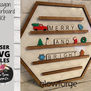 May include: A hexagon-shaped wooden letterboard kit with a white patterned background and brown frame. The words "MERRY AND BRIGHT" are spelled out with letter tiles. Decorative wooden Christmas-themed accents include a red truck, tree, and snowflake. The text "Hexagon Letterboard Kit" and "LASER SVG FILES" are also visible.