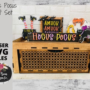 以下が含まれることがあります： 黒い猫、魔女の足、そして「Hocus Pocus」と「Amuck Amuck」と書かれた看板が付いた木製棚セット。