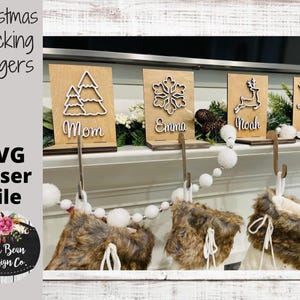 Puede incluir: Cuatro colgadores de calcetines de Navidad de madera con diseños recortados. Los diseños incluyen un copo de nieve, un reno, un árbol de Navidad y un trineo. Cada colgador tiene un nombre: Mamá, Emma, Noé y Papá.