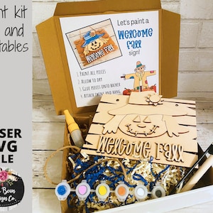 May include: A wooden craft kit to paint a welcome fall sign. The kit includes a wooden cutout of a scarecrow with the words "Welcome Fall" on it, paint, a paintbrush, and instructions. The kit is in a white box with a brown lid.