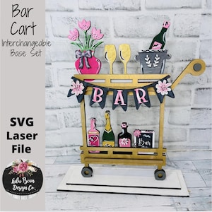 May include: A wooden bar cart with interchangeable decorations. The cart features a banner that spells out "BAR", a vase of pink flowers, champagne glasses, and bottles. The cart is on a white base and has the text "Bar Cart Interchangeable Base Set".