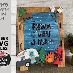 May include: A wooden camping sandwich board set with a blue background and the text "home is where we park it". The board features a camper, trees, a campfire, and a lantern.