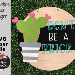 May include: A round wooden door hanger with a black and white striped background. The text "Don't be a prick" is written in turquoise lettering. A green cactus with pink hearts is on the left side of the hanger.