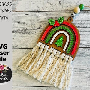 May include: A wooden Christmas rainbow ornament with a green Christmas tree and red holly berries. The rainbow has a white center and is layered with red and green stripes. The ornament has a white macrame tassel.