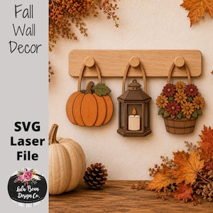 May include: A wooden wall rack with three hooks displaying hanging decorations: an orange pumpkin, a brown lantern with a white candle, and a basket of orange and red flowers. A real pumpkin and a pinecone sit on the surface below the rack.