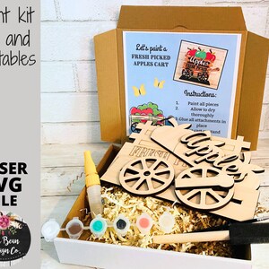 May include: A paint kit for a wooden craft project featuring a fresh picked apples cart. The kit includes wooden pieces, paint, brushes, and instructions. Let's paint a fresh picked apples cart.