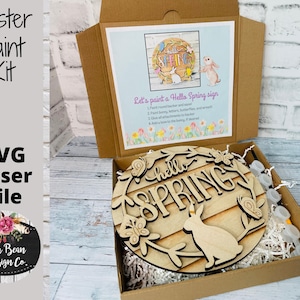 Può includere: Un kit di pittura pasquale in una scatola di cartone. Il kit include un cartello in legno con le parole "hello SPRING" e un disegno di coniglietto. La scatola contiene anche istruzioni ed elementi decorativi. Il kit è di Lulu Bean Design Co.