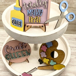 May include: A two-tiered white tiered tray with craft supplies. The top tier has a pink sign that says "I can totally make that" with a yellow glue bottle and blue scissors. The bottom tier has a pink sign that says "Craft Queen", a wooden paint palette with colorful paint blobs, and a pink glue gun.