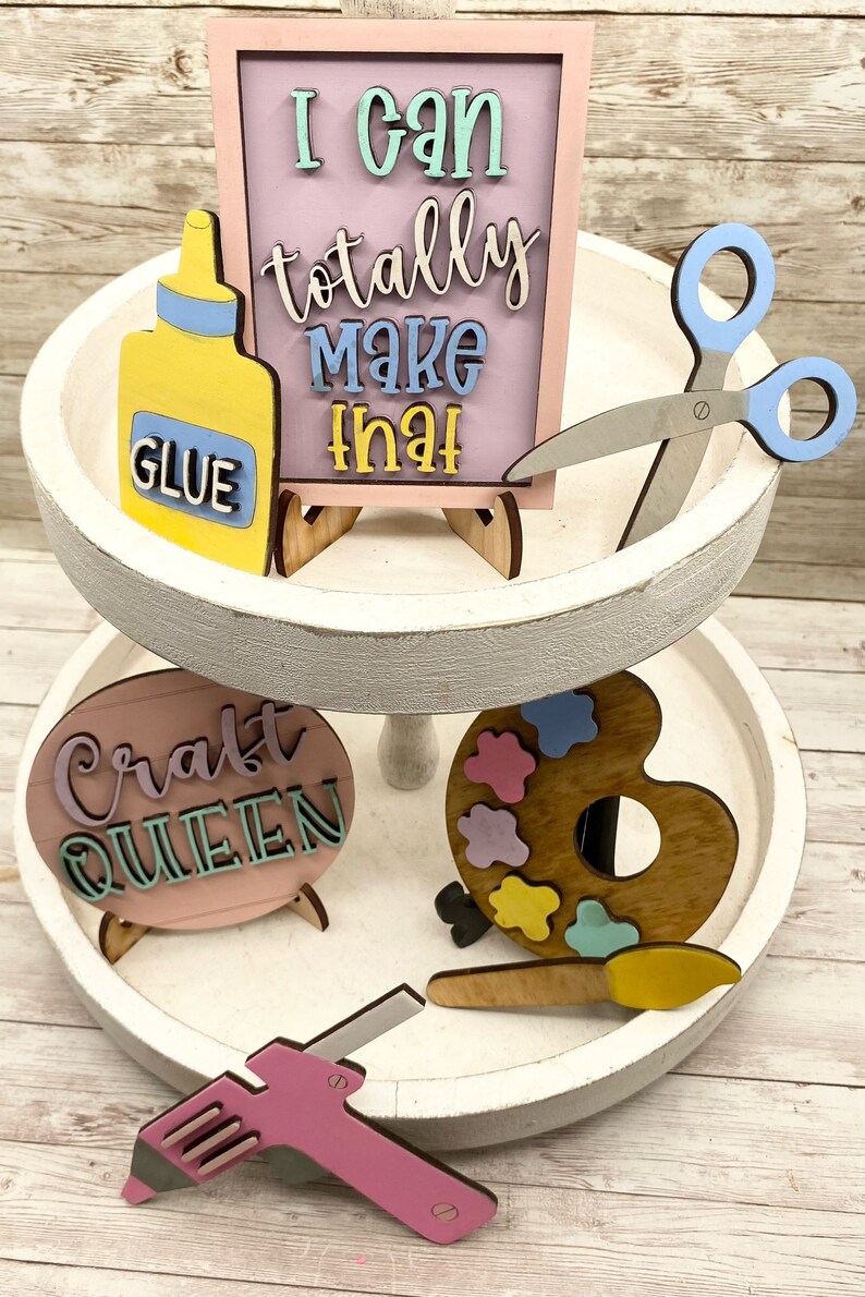 May include: A two-tiered white tiered tray with craft supplies. The top tier has a pink sign that says "I can totally make that" with a yellow glue bottle and blue scissors. The bottom tier has a pink sign that says "Craft Queen", a wooden paint palette with colorful paint blobs, and a pink glue gun.