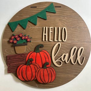 Puede incluir: Un letrero redondo de madera con las palabras "Hello Fall" en letras marrones. El letrero est&aacute; decorado con una pancarta de papel verde, una planta en maceta con flores rojas y tres calabazas naranjas.