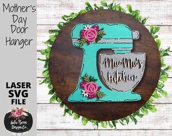 May include: A teal and gray wooden cutout of a stand mixer with a bowl that says "Mom-Mom's Kitchen". The mixer is decorated with pink and white flowers. The cutout is on a brown wooden background.