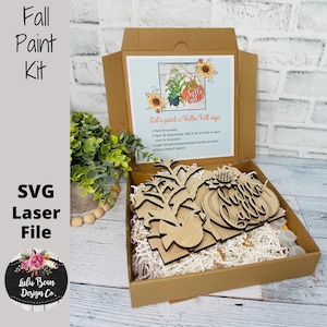 May include: A fall-themed paint kit in a cardboard box. The kit includes wooden cutouts of pumpkins, leaves, and a bird, along with instructions. The box also contains the text "Fall Paint Kit" and "SVG Laser File."