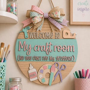 May include: A round wooden sign with the text "WELCOME TO My craft room (NO YOU CAN'T USE MY SCISSORS)". The sign has a large bow at the top and craft-themed embellishments. The sign is hanging on a wall with other craft supplies.