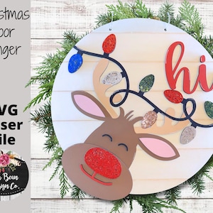 Puede incluir: Un colgador de puerta navideño redondo con un diseño de reno, una nariz roja y una cadena de luces de colores. La palabra "hi" está escrita en rojo. El diseño está sobre un fondo de vegetación y una superficie de tablones de madera.