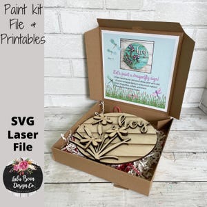 May include: A wooden craft kit featuring a round wooden sign that reads "hey" with dragonfly and flower cut-outs. The kit includes a printed instruction sheet with the text "Let's paint a dragonfly sign!" and a list of instructions.