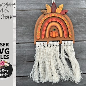 May include: A wooden Thanksgiving rainbow car charm with a pumpkin in the center. The rainbow is decorated with orange, red, and yellow paint and has a brown woodgrain finish. The charm has a white tassel hanging from the bottom.