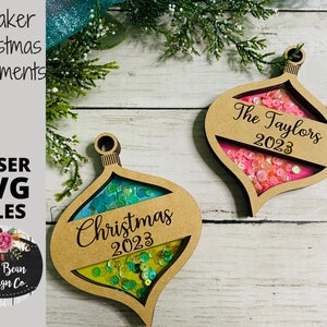 Może przedstawiać: Dwa drewniane kształty ozdób choinkowych z okienkiem do potrząsania. Ozdoby są ozdobione brokatem i tekstem "Christmas 2023" i "The Taylors 2023".