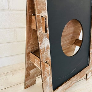Op de afbeelding: Een houten A-frame krijtbord met een verweerde witte verf. Het bord heeft drie planken en een ronde uitsparing in het krijtbord.