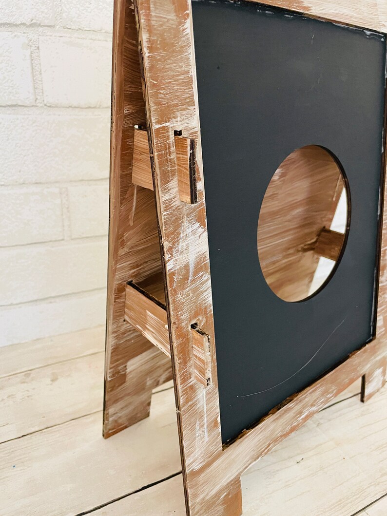 Op de afbeelding: Een houten A-frame krijtbord met een verweerde witte verf. Het bord heeft drie planken en een ronde uitsparing in het krijtbord.