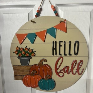 Puede incluir: Un colgador de puerta redondo de madera con fondo beige. Presenta una pancarta con tri&aacute;ngulos naranja y turquesa, una planta en maceta con flores naranjas, tres calabazas y el texto "Hello Fall" en letras rojas y negras.