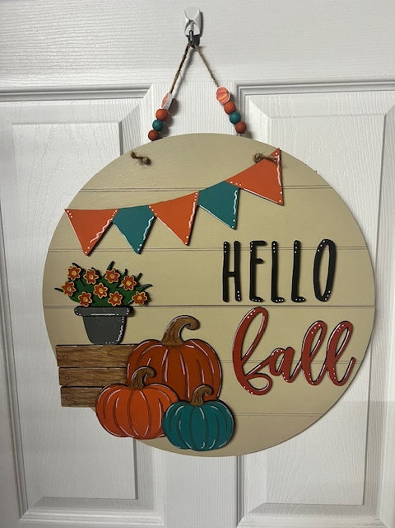 Puede incluir: Un colgador de puerta redondo de madera con fondo beige. Presenta una pancarta con tri&aacute;ngulos naranja y turquesa, una planta en maceta con flores naranjas, tres calabazas y el texto "Hello Fall" en letras rojas y negras.