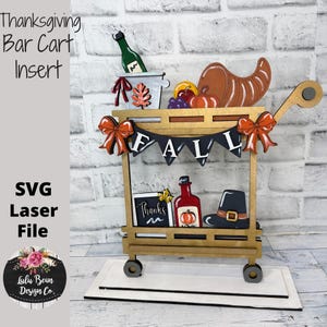May include: A wooden Thanksgiving bar cart insert with a banner that says "FALL". The cart is decorated with pumpkins, a cornucopia, and a bottle of wine. The cart has wheels and a handle. The text "SVG Laser File" is on the side.