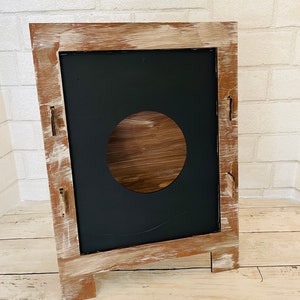 Op de afbeelding: Een rustiek houten bord met een verweerde witte verf afwerking. Het bord heeft een zwarte schoolbord inzet met een ronde uitsparing in het midden. Het bord staat op een houten ezel.