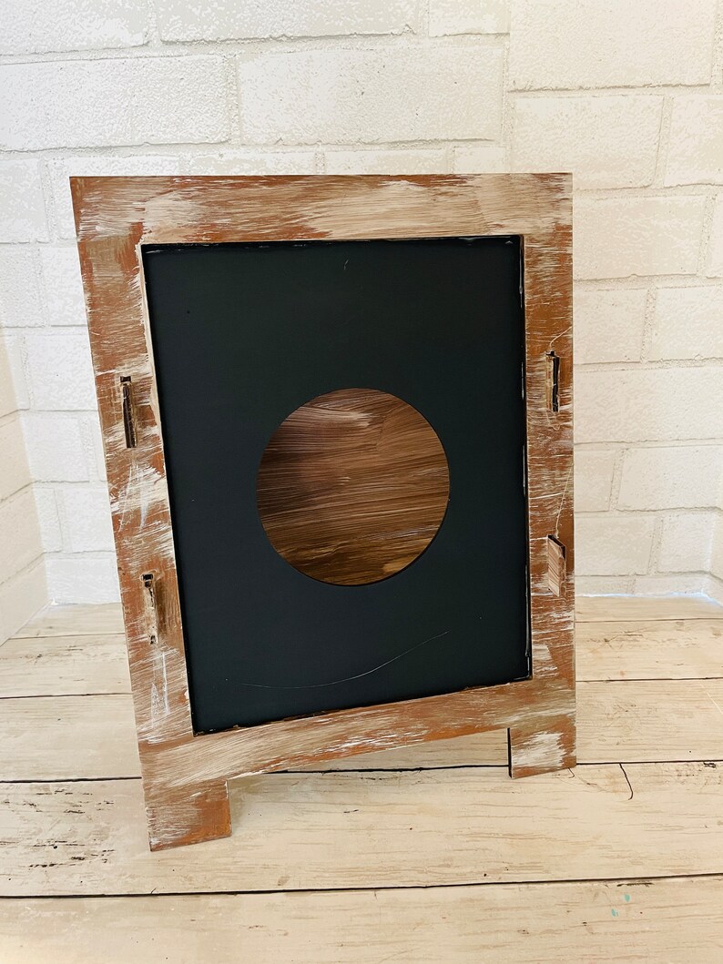 Op de afbeelding: Een rustiek houten bord met een verweerde witte verf afwerking. Het bord heeft een zwarte schoolbord inzet met een ronde uitsparing in het midden. Het bord staat op een houten ezel.