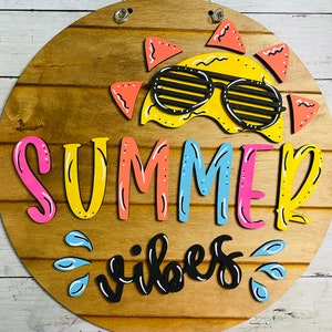 May include: A round wooden sign with the words "Summer Vibes" in colorful letters. A yellow sun with sunglasses is at the top of the sign.